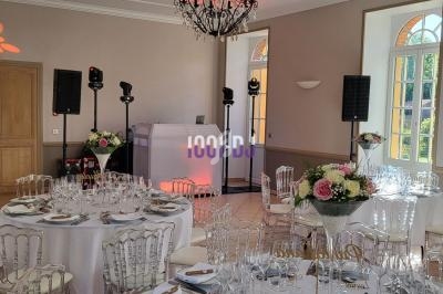 Salle de réception décorée avec des tables rondes dressées, un espace DJ équipé et des fleurs en centre de table.