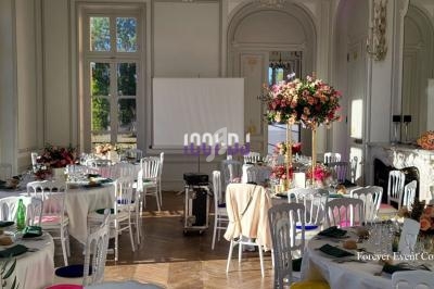 Salle de réception lumineuse avec tables décorées de fleurs colorées, chaises blanches et écran de projection.
