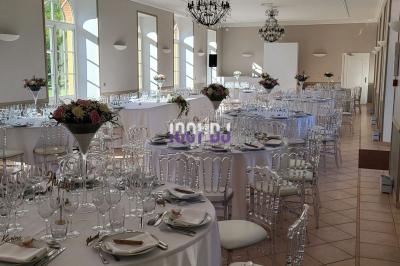 Salle de réception lumineuse avec tables rondes décorées de fleurs, vaisselle élégante et chaises transparentes.