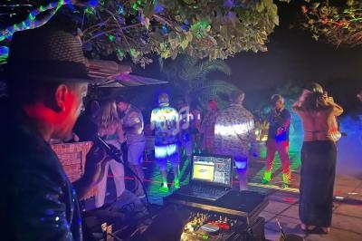 Un DJ anime une soirée en plein air avec des jeux de lumière colorés et des invités dansant sous des arbres.