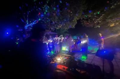 Personnes dansant en extérieur la nuit sous des lumières colorées, avec un DJ et son équipement au premier plan.
