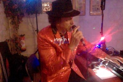 Un homme en veste rouge scintillante et chapeau noir chante au micro dans une pièce éclairée par des lumières colorées.