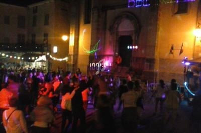 Foule dansant de nuit devant une église illuminée, avec des guirlandes lumineuses et des projections colorées.