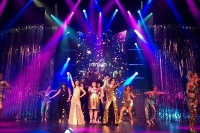 Des artistes en costumes brillants dansent et chantent sur une scène éclairée par des projecteurs colorés.