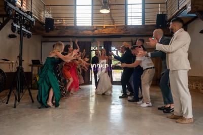 Un groupe de personnes applaudissant et dansant autour d'une mariée dans une salle lumineuse au style rustique.