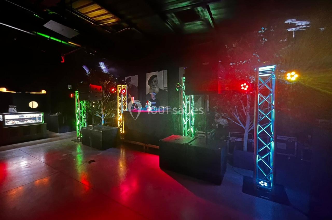 Scène de DJ dans une salle sombre avec éclairages colorés et structures lumineuses verticales.