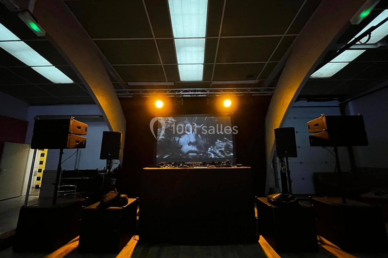 Salle sombre avec une scène équipée de platines DJ, des enceintes et un écran projetant un visage en noir et blanc.