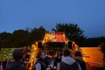 Un DJ joue de la musique sur une scène éclairée en soirée, devant un public rassemblé en plein air.