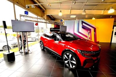Voiture électrique rouge exposée dans un showroom moderne, éclairée par des projecteurs avec écrans et décor coloré en…