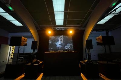 Salle sombre avec une scène équipée de platines DJ, enceintes et un écran projetant une image en noir et blanc.