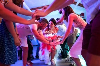 Des personnes dansent en cercle, une femme en robe blanche et rouge passe au centre sous un tunnel de bras.