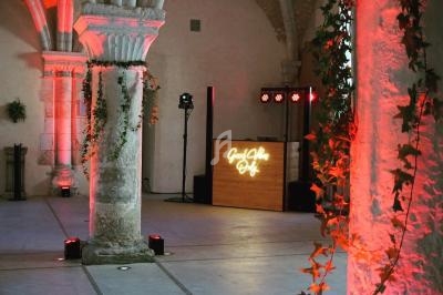 Salle voûtée avec colonnes en pierre, décorée de guirlandes végétales et éclairée par des lumières rouges et un DJ en…