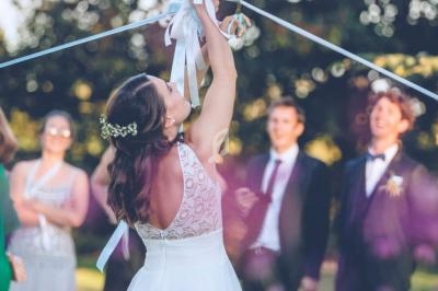 Une mariée en robe blanche tient un bouquet décoré de rubans, entourée d'invités dans un jardin.