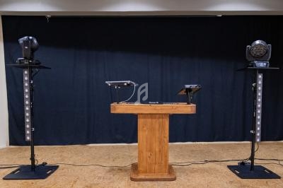 Un pupitre en bois entouré de deux colonnes sonores et projecteurs sur pied, dans une salle avec rideau noir.