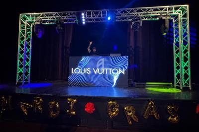 Un DJ mixe sur une scène éclairée par des lumières colorées, avec un écran affichant ’Louis Vuitton’.