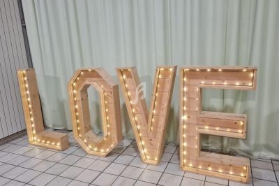Lettres en bois formant le mot ’LOVE’, illuminées par des guirlandes lumineuses, posées sur un sol carrelé devant un rideau…