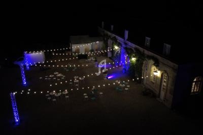 Cour extérieure éclairée de nuit avec guirlandes lumineuses, tables rondes et bâtiment ancien en arrière-plan.