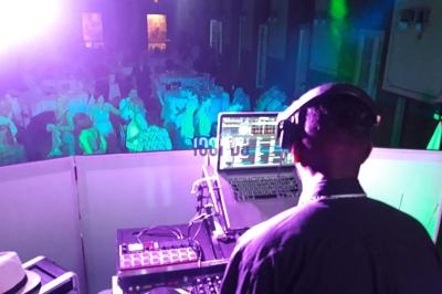 Un DJ de dos mixe sur une console, face à une salle éclairée de lumières colorées avec des invités assis.