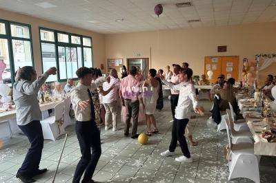 Des personnes dansent dans une salle décorée pour une fête, entourées de tables dressées et de ballons au sol.