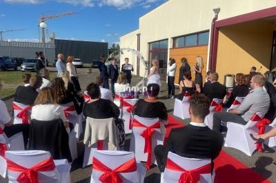 Cérémonie en extérieur avec des invités assis sur des chaises blanches décorées de nœuds rouges, près d'un bâtiment.
