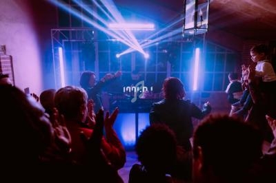 Groupe de personnes applaudissant et dansant devant un DJ sous des lumières colorées dans une salle sombre.