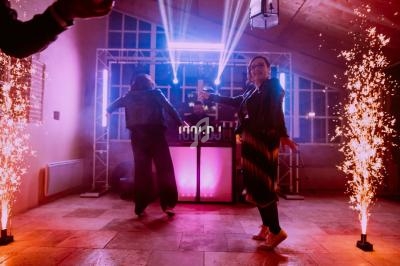 Deux personnes dansent devant un DJ avec des lumières colorées et des étincelles sur les côtés.