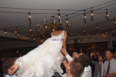 Une mariée en robe blanche est soulevée par des invités sous des guirlandes lumineuses dans une salle de réception.