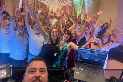 Un groupe de personnes souriantes levant les bras lors d'une fête, avec un DJ au premier plan et une ambiance festive.