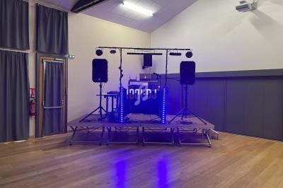 Installation de matériel de DJ sur une estrade avec éclairage LED bleu dans une salle aux murs clairs et rideaux gris.