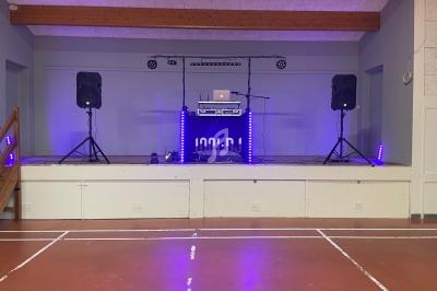 Scène avec équipement de sonorisation, lumières violettes, deux enceintes, et un ordinateur portable sur une table.