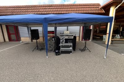 Matériel de sonorisation installé sous un barnum bleu, comprenant enceintes, table de mixage et câblage.