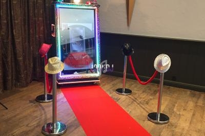 Miroir interactif entouré de chapeaux et d’accessoires, avec un tapis rouge et des cordons de séparation.