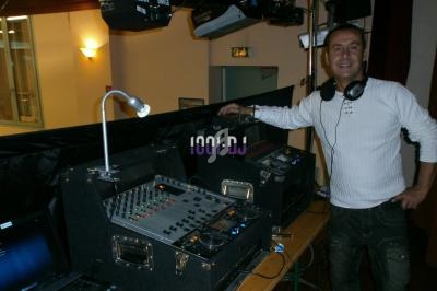 Un homme souriant debout près d'une table de mixage et d'équipements audio dans une salle éclairée.