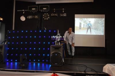 Un homme souriant se tient près d'une scène équipée de matériel audio et lumineux, avec une projection vidéo en arrière-plan.