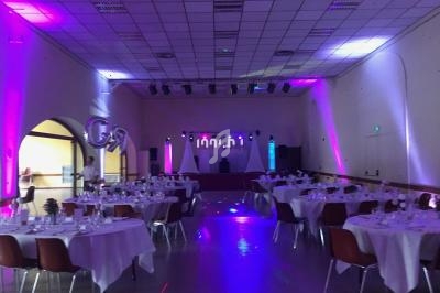 Salle de réception décorée avec des tables dressées, éclairages colorés et une scène équipée pour un événement.