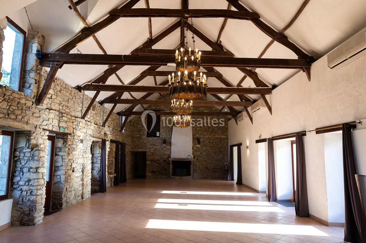 Salle spacieuse avec murs en pierre, poutres apparentes, lustres suspendus et grandes fenêtres laissant entrer la lumière.