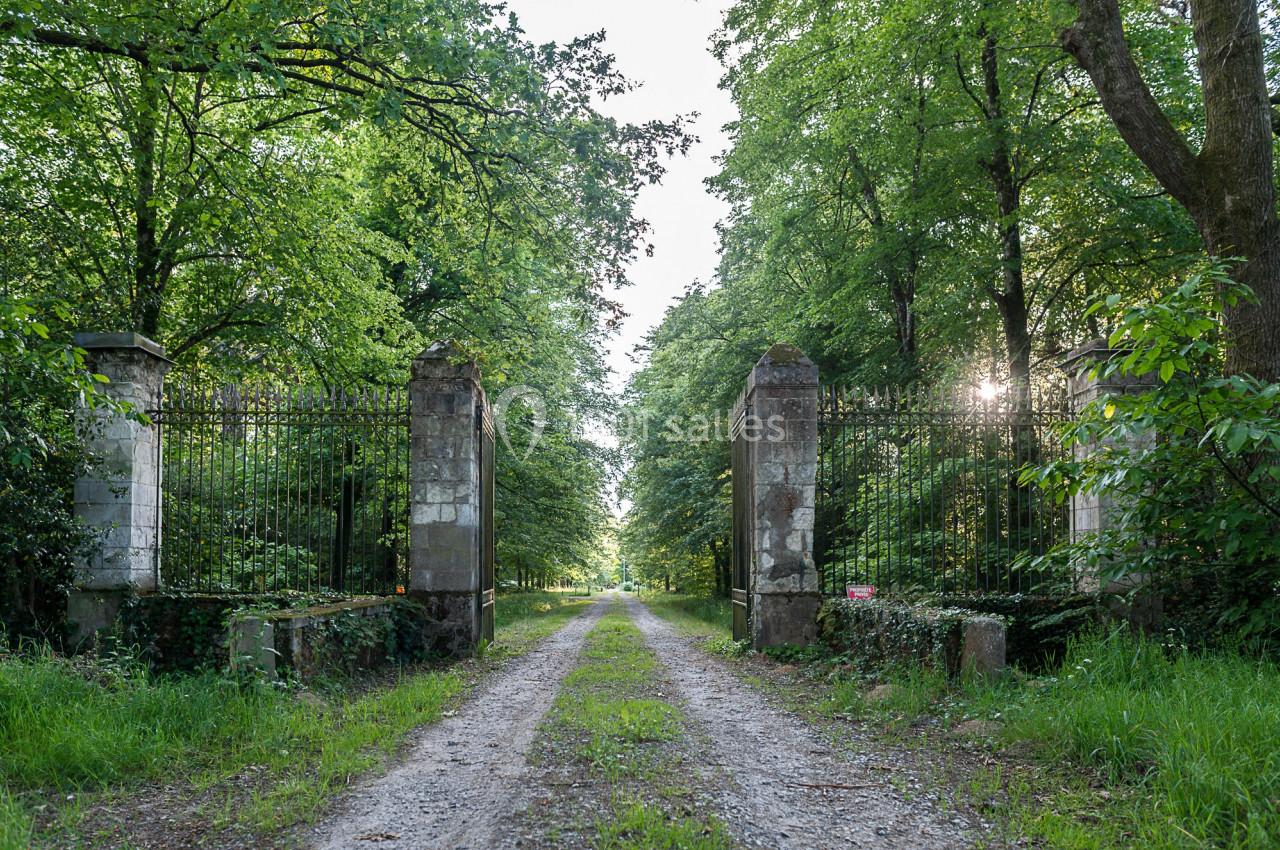 Chemin de terre bordé d'herbe menant à une allée arborée, encadré par un portail en fer forgé et des piliers en pierre.