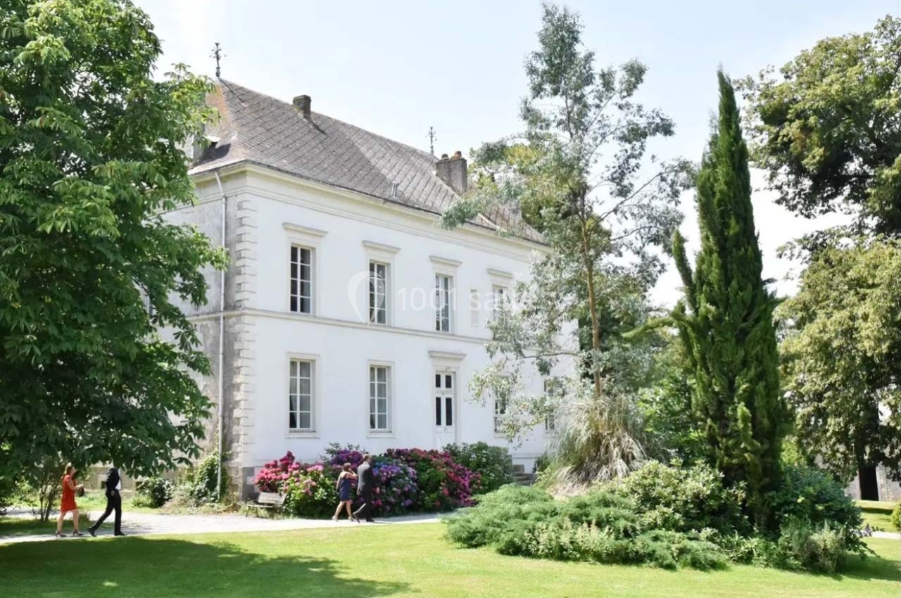 Grande maison blanche entourée de jardins verdoyants avec des arbres, des arbustes et des fleurs colorées.