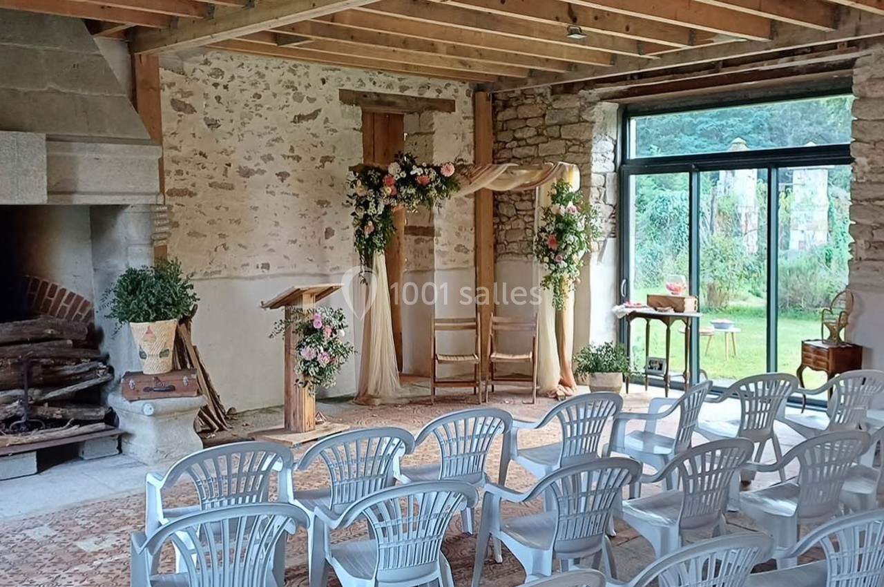 Salle de cérémonie avec chaises blanches, arche fleurie, cheminée en pierre et vue sur un jardin par une baie vitrée.
