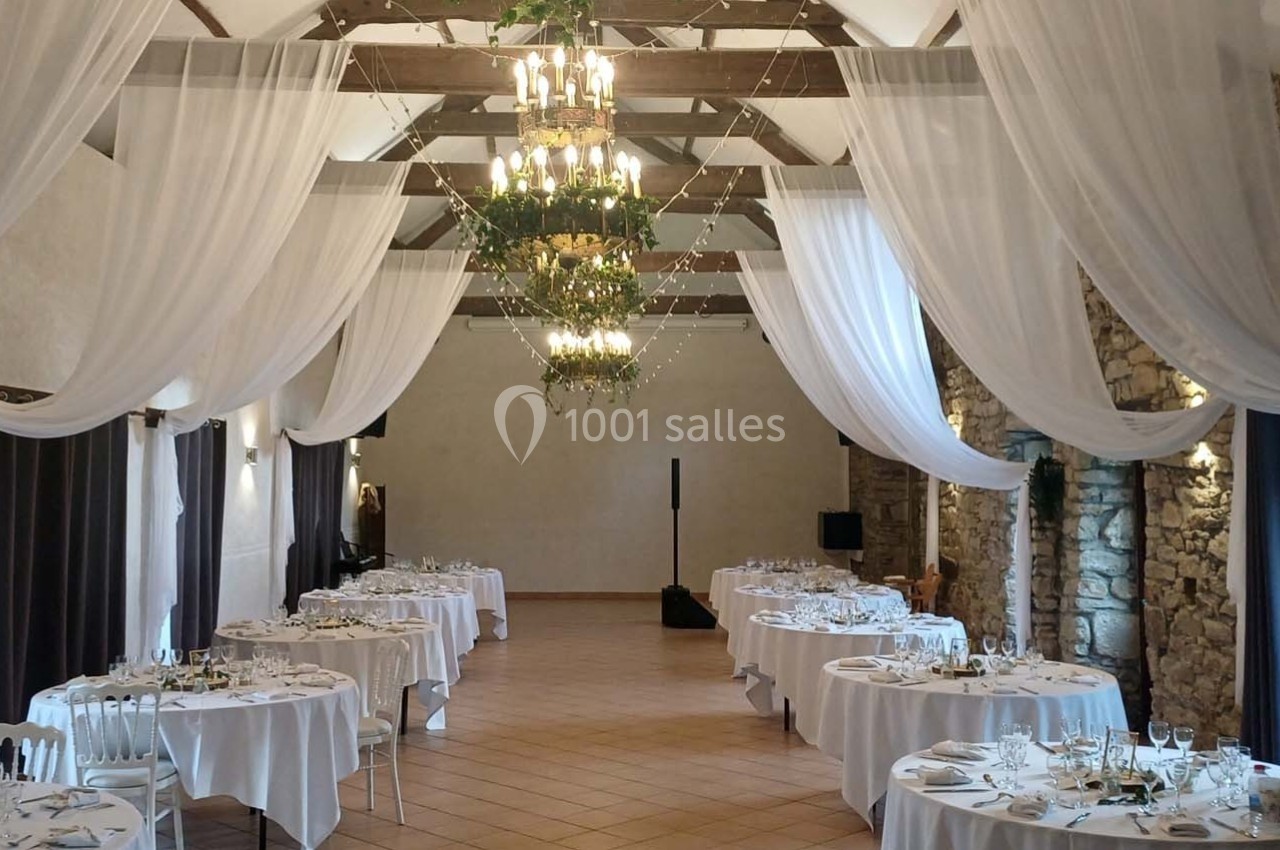 Salle de réception avec tables rondes dressées, rideaux blancs suspendus et murs en pierre apparente.
