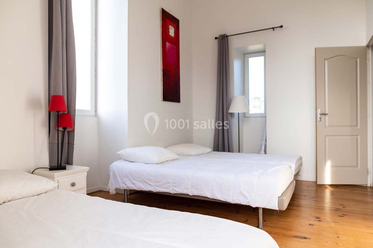 Deux lits simples dans une chambre lumineuse avec parquet, rideaux gris, lampe rouge et tableau mural.