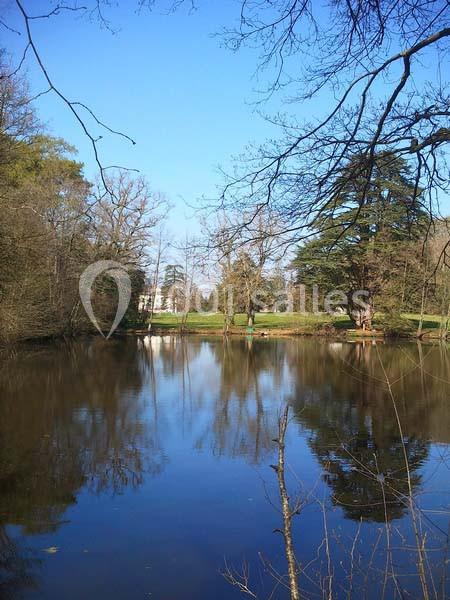 Étang calme entouré d'arbres avec un bâtiment blanc visible en arrière-plan sous un ciel bleu.