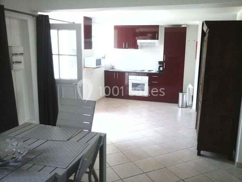 Cuisine ouverte avec meubles rouges, table à manger en métal gris et sol en carrelage beige.