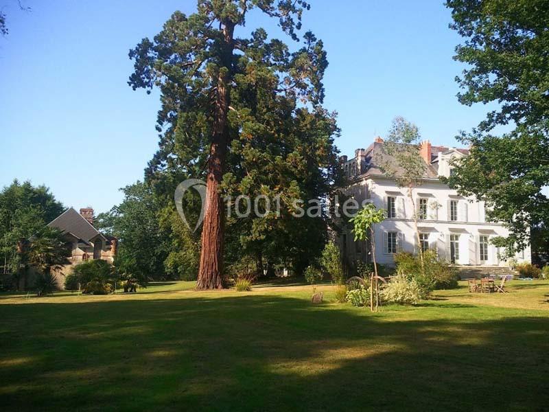 Grande maison blanche entourée d'un jardin verdoyant avec un grand arbre au premier plan, sous un ciel dégagé.
