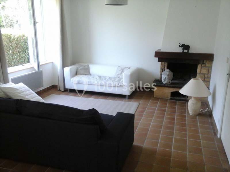 Salon avec canapé blanc et noir, cheminée en brique, lampe et décoration, sol en carrelage et fenêtre ouverte sur jardin.