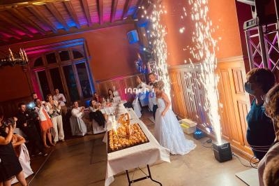 Un couple de mariés coupe leur pièce montée dans une salle décorée, entouré d'invités et d'effets pyrotechniques.