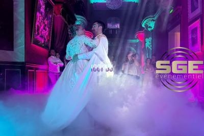 Un couple danse dans une salle éclairée de lumières colorées, entourée de fumée et d'invités en arrière-plan.