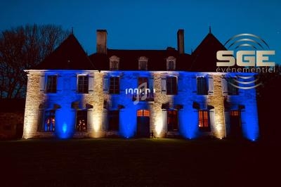 Façade d'un bâtiment ancien éclairée en bleu la nuit, avec un logo ’SGE événementiel’ superposé à droite.