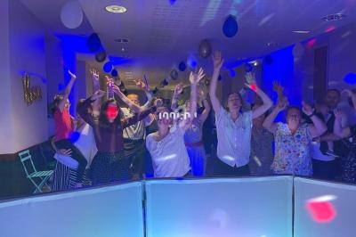 Des personnes dansent et lèvent les bras sous des lumières colorées dans une salle décorée de ballons.