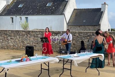 Deux musiciens jouent en plein air devant un mur en pierre, avec des invités discutant près de tables dressées.
